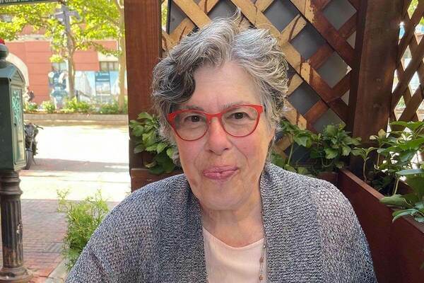 Helen Epstein is a woman with grey and black hair. She is wearing red glasses and a grey cardigan. Behind her is a lattice separating the interior from the street outside. Outside is clearly a bright and sunny day, and a street and a tree are brightly lit.