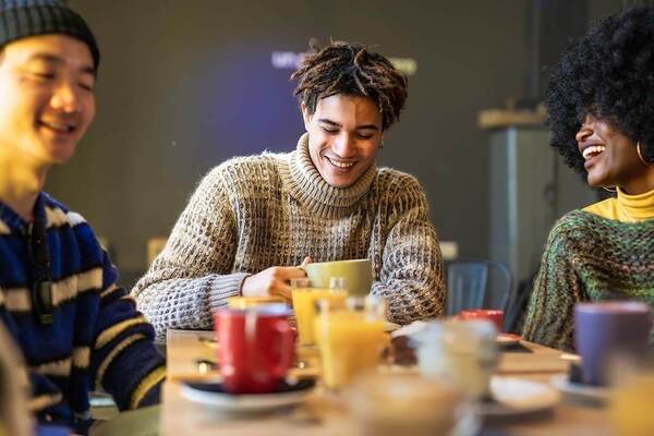 Three students wearing cozy sweaters share a laugh and refreshments around a table. The focus is on a smiling student with textured hair, holding a mug. Various mugs and glasses of orange juice are on the table.