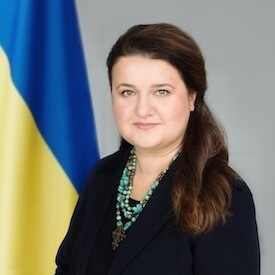 Headshot of Ambassador Oksana Markarova, who is wearing a green beaded necklace and navy blazer. She is sitting in front of a Ukrainian flag.