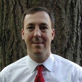 Outdoors headshot of Carl Bruch, who is wearing a white button-up shirt and a red tie.