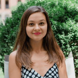 Outdoors headshot of Anna Romandash, who is standing in front of a large green shrub and is wearing a black dress with polka dots on it.