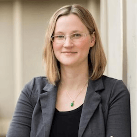 Headshot of Sarah Wilson Sokhey, who is wearing thin glasses, a teal/gold necklace, a black shirt, and a grey blazer.