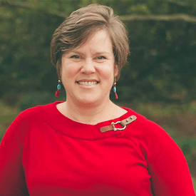 Outdoors headshot of Sarah D. Phillips, who is wearing teal/red earrings and a red sweater.