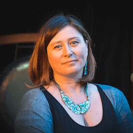Headshot of Halyna Kruk, who is wearing a teal mosaic necklace and matching earrings. She is also wearing a black shirt and grey sweater.