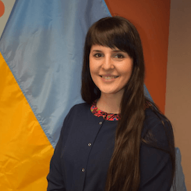 Headshot of Svitlana Ramer, who is wearing a navy sweater and standing in front of the Ukrainian flag.