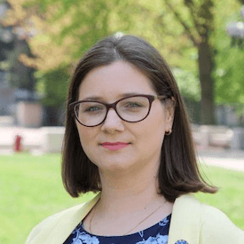 Outdoors headshot of Katerina Sirinyok-Dolgaryova, who is wearing brown glasses, a yellow blazer, and blue floral shirt.
