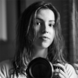 This is a photo of Iva Sidash. It is a unique style of headshot, as it is in black and white. Her camera is in the foreground and in focus, while she is slightly out of focus behind it.