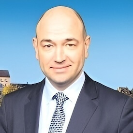 A man wearing a dark suit and patterned tie smiles against a blurred background of buildings and blue sky.