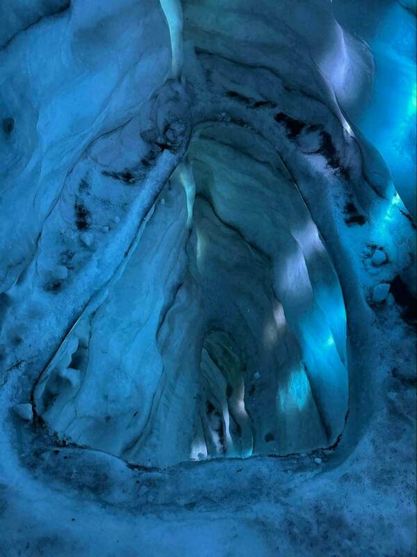 Looking down into a glacial crevasse; vibrant blue light emanates from deeper within the ice, illuminating the textured walls.