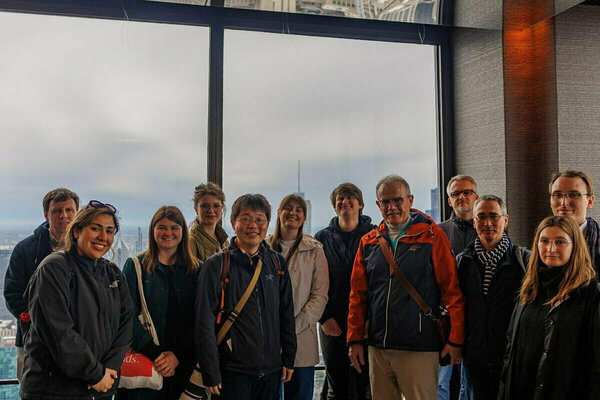 A group of eleven people stands before large windows offering a cloudy cityscape view.