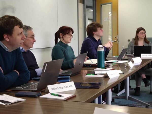 Five people sit at a long table during a meeting. They have laptops open and name placards in front of them.  A whiteboard is visible in the background.