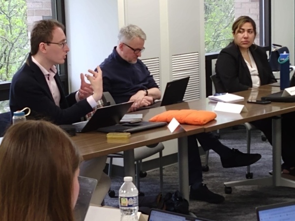 Three people sit at a conference table with laptops. A man in a dark jacket and pink shirt gestures while speaking.  Another man in a blue sweater types on his laptop. A woman in a black jacket listens.
