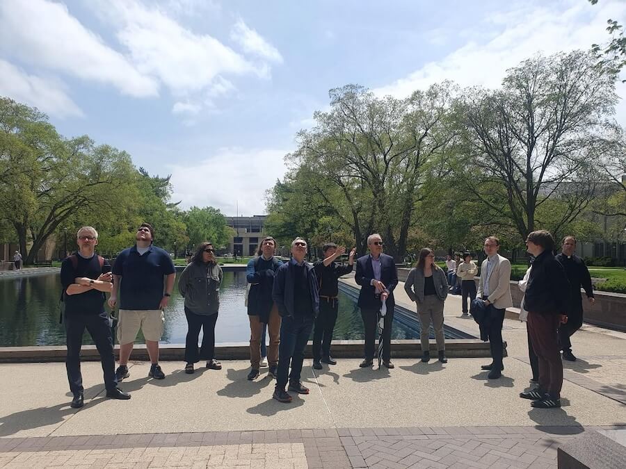 A group of people stands by the reflecting pool outside the Hesburgh Library on the University of Notre Dame campus, some looking upwards.