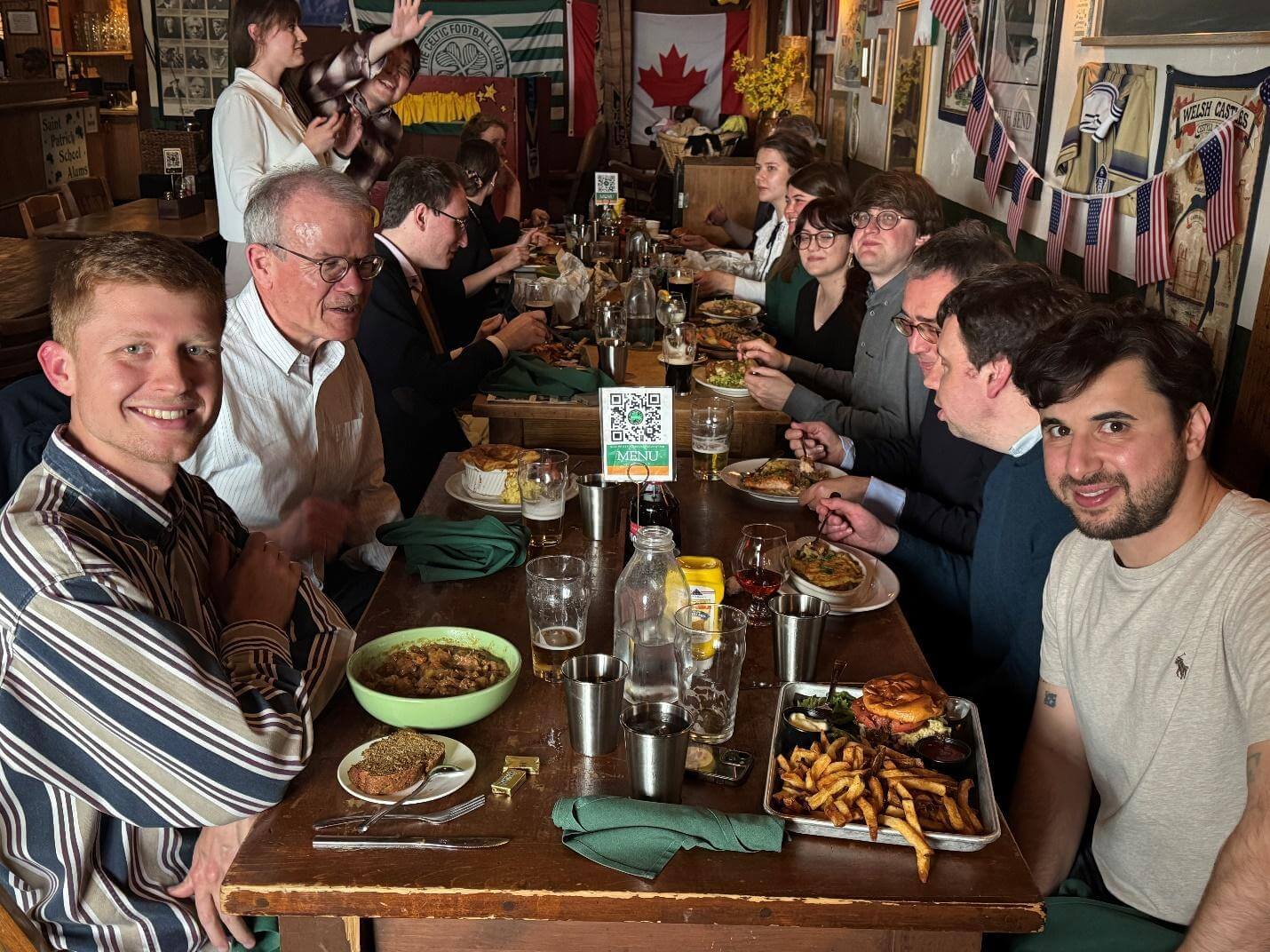A group of people sit at a long wooden table, enjoying a meal and conversation in a pub decorated with Notre Dame and Celtic memorabilia.