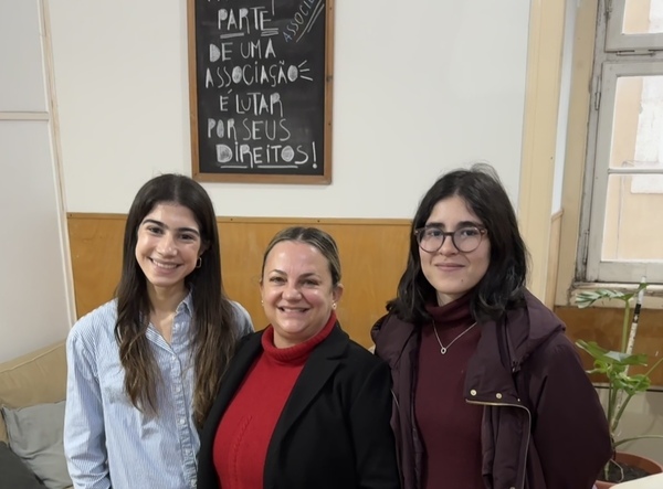 Three women stand smiling in front of a chalkboard that reads, in Portuguese, "Join an association and fight for your rights!" in white chalk.  The woman on the left wears a light blue and white striped shirt. The woman in the center wears a red turtleneck sweater and black jacket.  The woman on the right wears glasses and a dark maroon jacket.