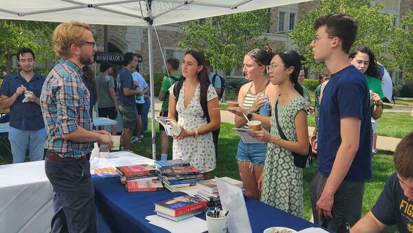Students talk with a man at a table with books under a white tent on the University of Notre Dame campus.