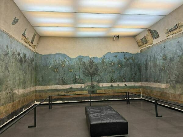 Restored Roman fresco depicting a garden scene with trees, birds, and a distant mountain range in blues and greens, displayed in a museum setting with a simple black bench in the foreground.