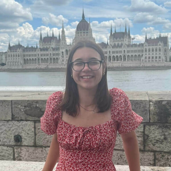 A young woman with medium-length brown hair and a red dress, Sofia Buck, stands in front of the Parliament of Budapest.