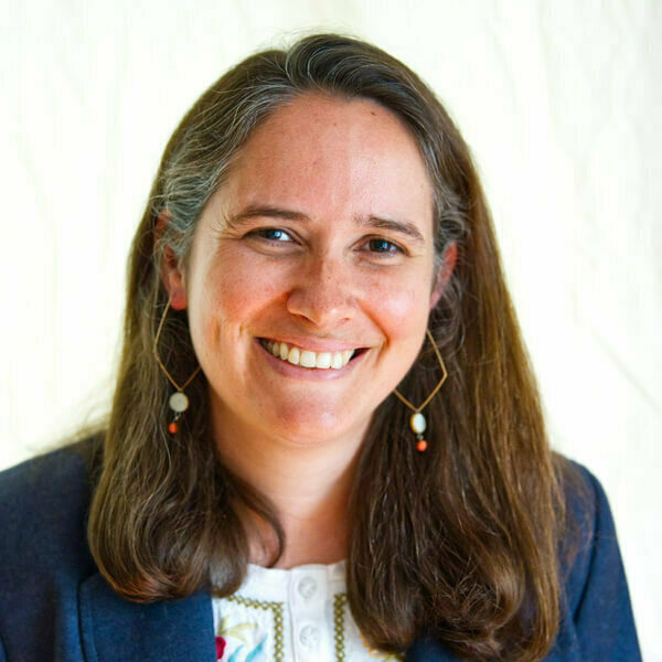 A woman with brown hair, a blue sweater, and fun, geometric earrings, Rebecca Kates, smiles in front of a white background.