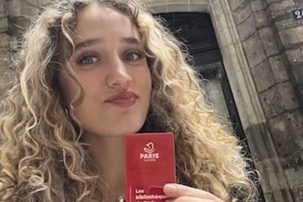 A young woman with light brown curly hair holds up a red 'Les bibliothèques de Paris' library card, making a puckered face. She stands before the ornate stone entrance of Hôtel de Lamoignon, featuring sculpted cherubs.