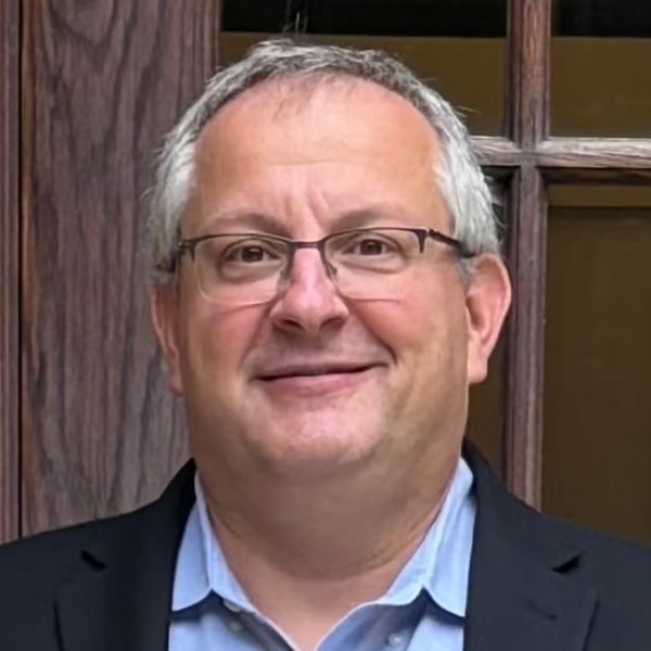 A man with graying hair and glasses smiles broadly, wearing a light blue shirt and dark jacket, in front of a dark wooden window frame.