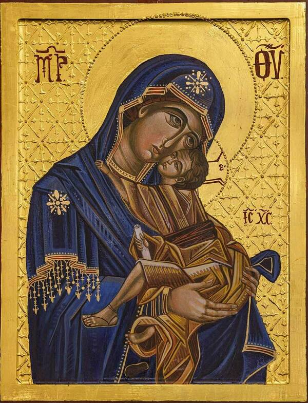 Icon depicting the Virgin Mary in blue, tenderly cradling the Christ Child. He wears a gold tunic, holds a scroll, and offers a blessing. Both have serious expressions against a rich, patterned gold background with red Greek text.