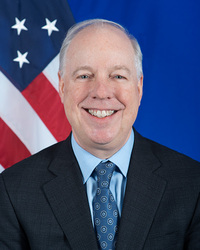 Kent Logsdon, a smiling man with light grey hair, wearing a dark suit, light blue shirt, and patterned blue tie. A US flag is visible behind him on the left.