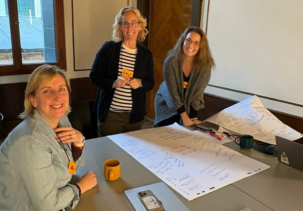 Three smiling women stand around a table with large white papers covered in blue handwritten brainstorming notes, diagrams, and circled words like "HOPE."