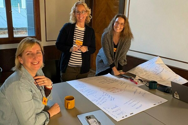 Three smiling women stand around a table with large white papers covered in blue handwritten brainstorming notes, diagrams, and circled words like "HOPE."