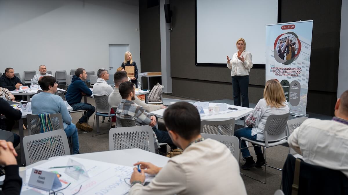 A woman in a light blouse speaks, gesturing to an audience of mostly men seated at tables in a presentation room. A banner promoting