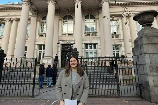 A smiling young woman in grey coat holds papers before a beige building with classical columns and iron gates.