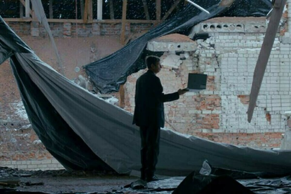 Man in a dark suit holds a laptop inside a severely damaged building with exposed brick, debris, and torn tarps.