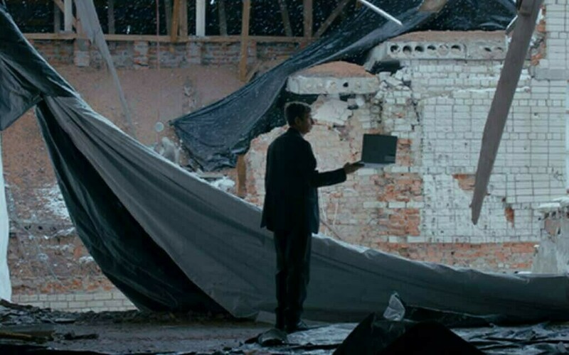 Man in a dark suit holds a laptop inside a severely damaged building with exposed brick, debris, and torn tarps.