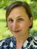 A woman with short light brown hair, green eyes, and a gentle smile wears a navy and white patterned shirt, green foliage background.