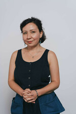 A woman with dark hair and a gentle smile wears a navy sleeveless top, colorful earrings, a necklace, and a bracelet. Her hands are clasped.