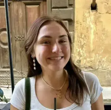 Smiling young woman with brown hair, white top, and pearl earrings. Historic building facade in soft focus background.