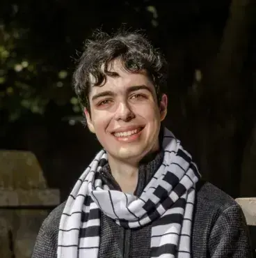 Young man with dark curly hair smiles, wearing a black and white checkered scarf and dark sweater.