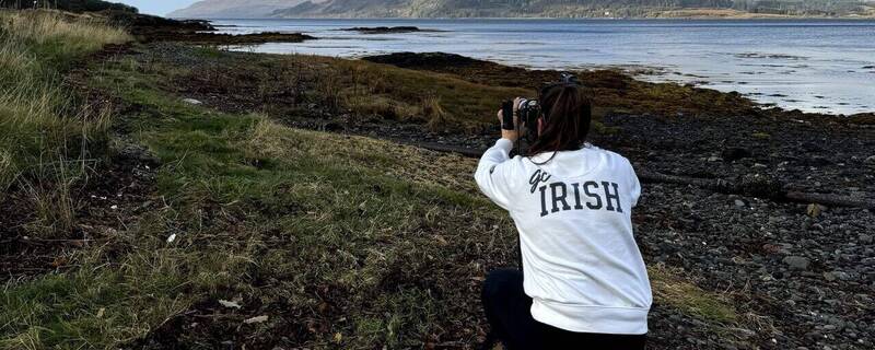 Person in white "go IRISH" sweatshirt crouched on a rocky shore, photographing blue water and distant hills under a cloudy sky.