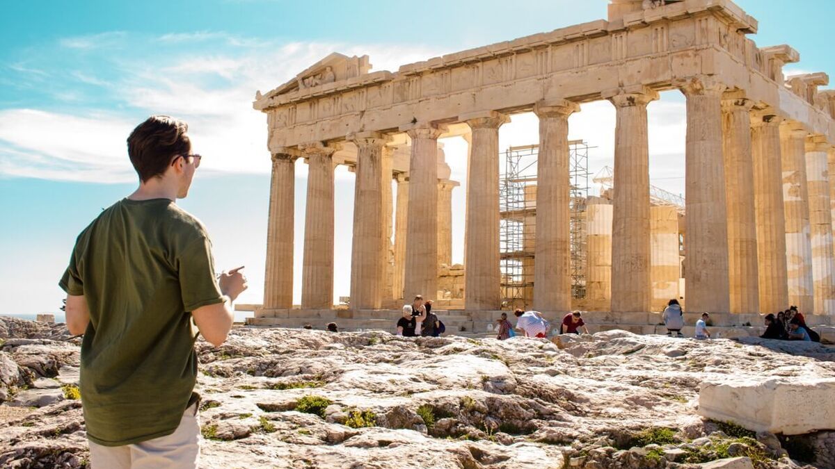 A man in a green shirt and sunglasses points towards the ancient Parthenon, sun-drenched with scaffolding.