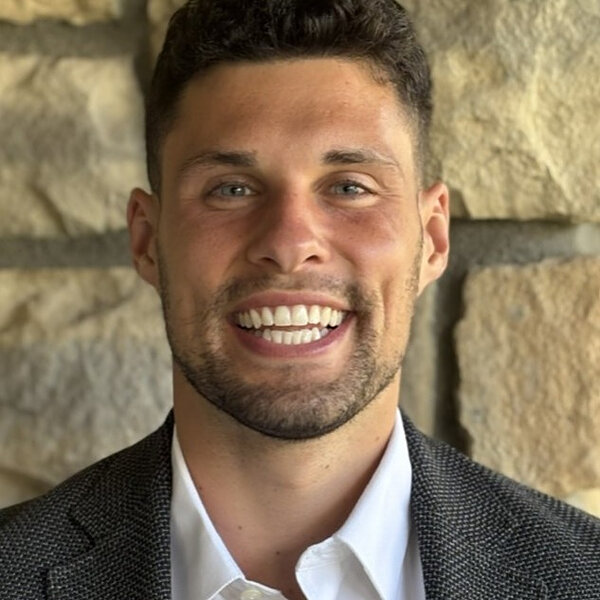 Nicholas Dawes smiles broadly, wearing a white dress shirt and a dark jacket.